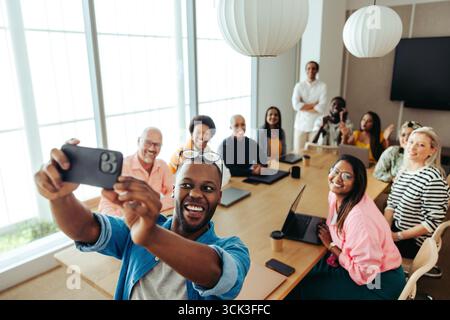 A diverse group of colleagues taking a joyful selfie in a modern office environment. The scene reflects a positive workplace culture where everyone is Stock Photo