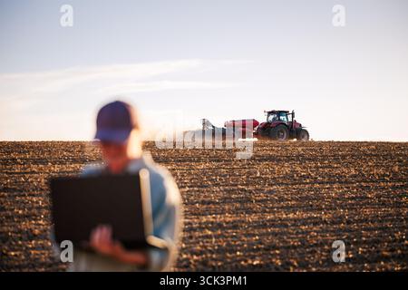 Smart farming and modern agriculture with farmer using laptop while tractor with seeder sowing crop seeds on field. Agronomist with computer analyzing Stock Photo