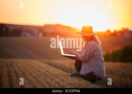 Woman agronomist analyzing smart farming data on laptop after harvest on agricultural field during sunset Stock Photo