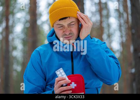 Hiker experiencing intense discomfort from a headache during a winter trek, reaching into a first aid kit for painkillers amidst a serene, snowy forest landscape Stock Photo