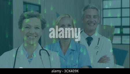 Standing three staff wearing lab coats, stethoscopes and scrubs in hospital corridor with signage Stock Photo