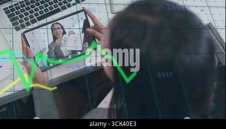Woman in gray blazer using tablet at desk showing laptop, document, notebook, pen, graphs on call Stock Photo