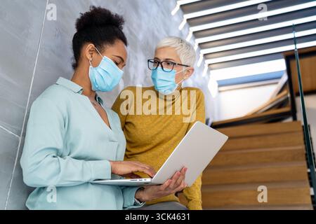 Female professionals in masks using laptop for technology, AI, digital innovation and safe workplace Stock Photo