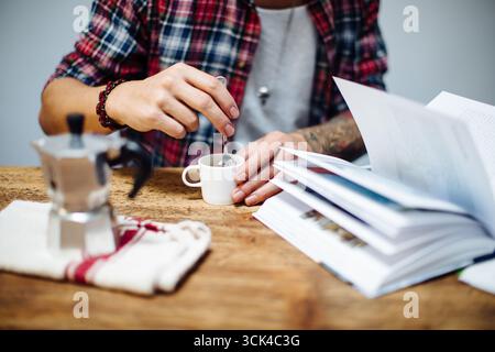 Young man stirring his coffee while reading a book Stock Photo