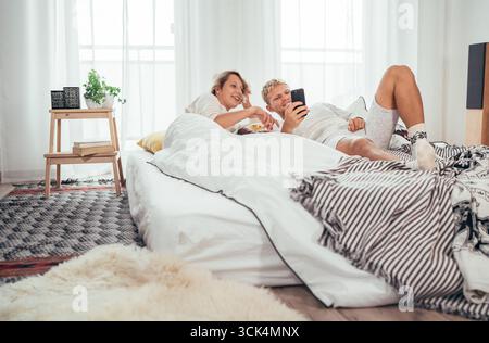 Young adults couple lying in cozy bed in bedroom and browsing together internet or checking mails using modern smartphone. Modern technology, and coup Stock Photo