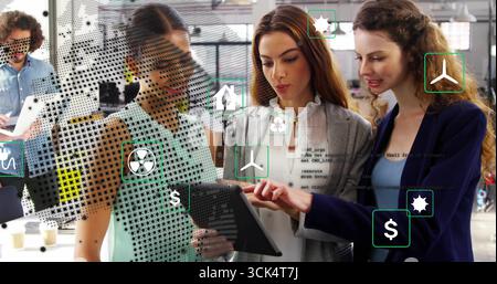 Team examining tablet in office featuring green icons, money icons, code icons, laptop, coffee cups Stock Photo