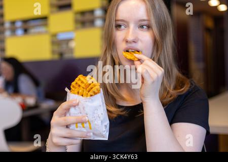 Kerpen, Germany - August, 9, 2025: A young woman takes a bite of waffle fries at a vibrant in McDonalds, modern eatery. She enjoys a casual lunch whil Stock Photo