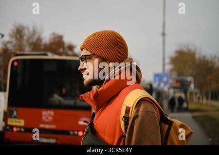 Side profile of a man in an orange beanie and glasses standing near a bus, dressed in a red vest and brown hoodie with a yellow backpack. Stock Photo