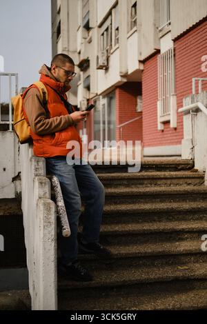 Young man leaning on stair railing and using smartphone, dressed in orange vest and jeans. Stock Photo