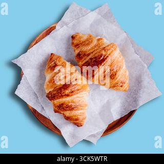 Two golden-brown croissants on parchment paper, served on a wooden plate, perfect for breakfast or a snack. Stock Photo