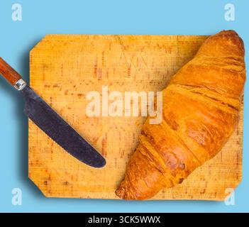 A croissant placed on a wooden cutting board next to a knife, ready for serving or preparation. Stock Photo