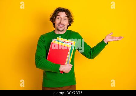 Young man in green pullover holding colorful notebooks gesturing hand sign on vibrant yellow background Stock Photo