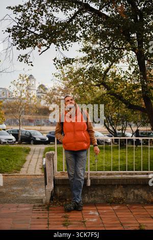 Man in orange vest standing under tree, holding glasses, with Belgrade city view in background. Autumn in town. Stock Photo