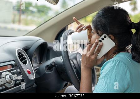 A woman is holding a smartphone to her ear while sipping coffee, driving in a city. Stock Photo