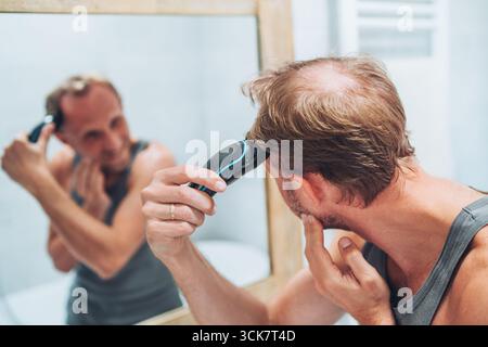 Body and skincare concept. Smiling man giving himself a new haircut using electric rechargeable trimmer while looking in bathroom mirror, personal gro Stock Photo
