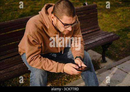 Man in glasses using smartphone on wooden bench in park, casual autumn outfit. Stock Photo