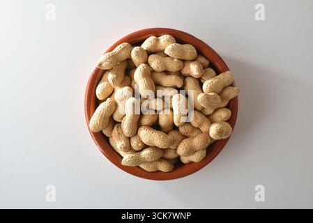 Heap of peanuts with skin in a brown clay bowl isolated on a white table. Top view. Stock Photo