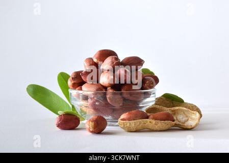 Heap of peeled peanuts with skin in a glass bowl isolated on a table with leaves and white isolated background. Front view. Stock Photo
