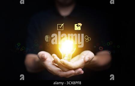 Bright light bulb in hands with brain and puzzle icons representing inspiration, leadership, innovation, teamwork, intelligence, and business solution Stock Photo