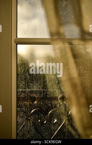 Sunlit raindrops on a windowpane with a soft glimpse of the window frame and sheer curtain, gently blurred. A warm, peaceful scene evoking quiet moments of calm after spring rain. Vertical photo. Stock Photo