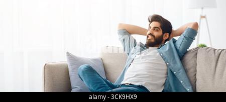 Home comfort. Young arab man relaxing on cozy sofa, leaning back on couch with hands behind head, free space Stock Photo