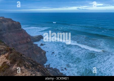 A dramatic coastal cliff overlooks powerful ocean waves crashing against rocky shores under a moody blue sky with scattered clouds on  Praia das Conch Stock Photo