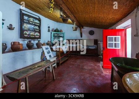 A traditional portuguese rustic kitchen with clay pots, wooden furniture, hanging gourds, and a bright red door under a thatched ceiling in Lourinhã Stock Photo