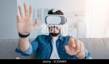 Future technology concept. Man playing video games wearing virtual reality glasses and gesturing, sitting on couch Stock Photo
