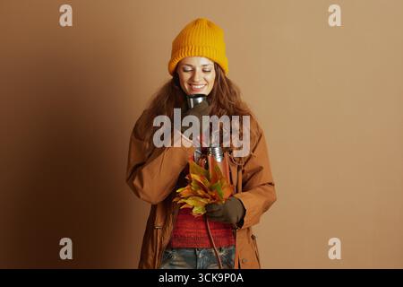 A woman in a yellow beanie and green gloves gently sips from a metal cup with closed eyes and a smile, holding autumn leaves. A thermos signifies warm Stock Photo