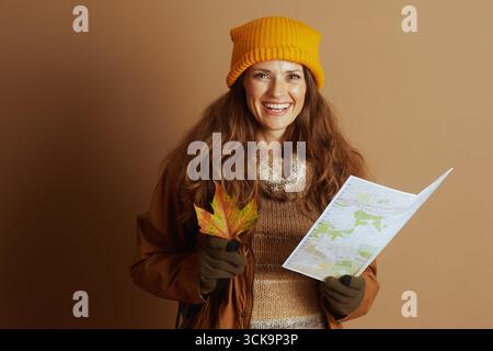 A smiling woman in autumn attire, wearing a yellow knit hat and brown jacket, holds a colorful fall leaf and a folded map, ready for a thoughtful outd Stock Photo