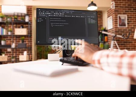 Closeup of computer screen showing programming scripts in progress in brick wall apartment. African American web developer seated at desk, writing code on desktop pc in brick wall home. Stock Photo
