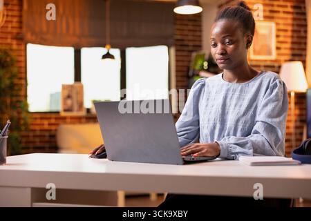 Young black woman uses laptop in cozy living room, managing tasks remotely. Focused female freelancer working on online projects, doing research and replying to emails from apartment. Stock Photo