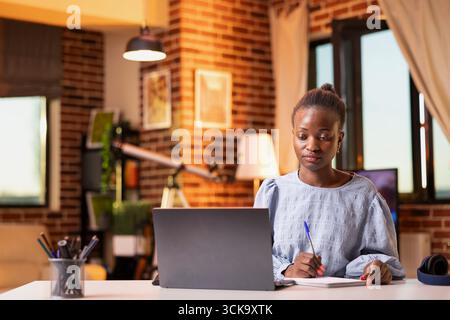 Confident businesswoman analyzing sales information and preparing reports from home. Female entrepreneur manages business tasks at desk with concentration, comparing data on laptop and notepad. Stock Photo
