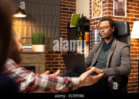 Caucasian man sharing relationship struggles during marriage counseling as focused therapist takes notes on laptop. Psychotherapy session emphasizes communication, mental wellbeing and trust. Stock Photo