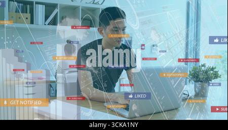 Leaning Asian man analyzing laptop screen in office with potted plant, floating notification icons Stock Photo