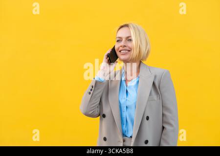 Young businesswoman in blazer with short blonde hair talking on smartphone standing against yellow background, smiling and looking confident Stock Photo