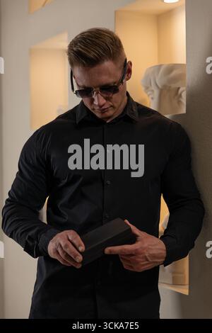 man wearing a fitted black shirt and sunglasses stands against a wall in a contemporary indoor area focused on a small black box he is holding. Soft lighting enhances the ambiance. Stock Photo
