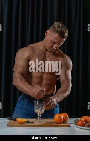 A muscular man in jeans squeezes an orange into a glass while standing in a kitchen. Fresh oranges are visible on the table showcasing a vibrant cooking scene. Stock Photo