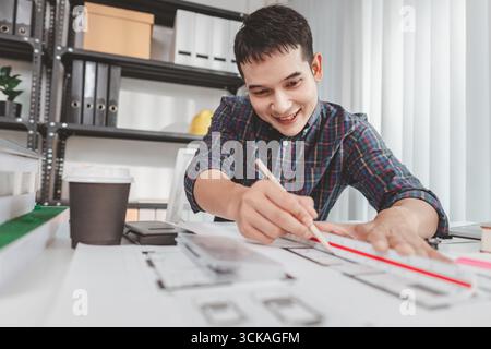 Young male architect working at office desk, drawing construction plans and using drafting tools, focused on building design while showing creativity Stock Photo