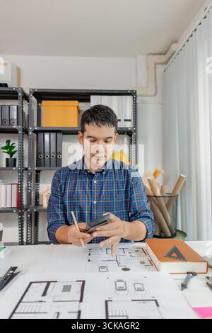 A young architect works on house design plans at his desk, focusing on drawings and scale models. The scene conveys creativity, dedication, and precis Stock Photo