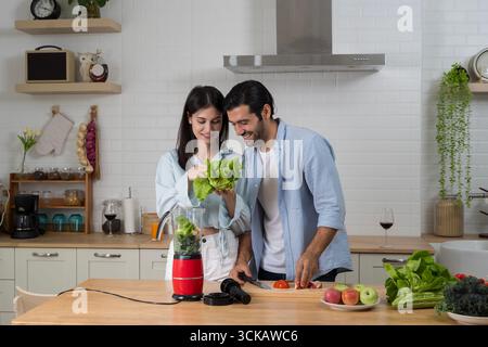 Smiling couple in kitchen preparing vegetables with blender for smoothie, promoting healthy lifestyle, nutrition, organic food, teamwork, wellness, co Stock Photo
