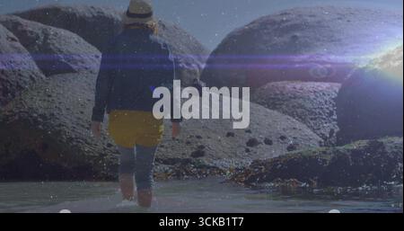 Wading woman wearing boots and straw hat traversing shallow ocean water, with boulders and seaweed Stock Photo