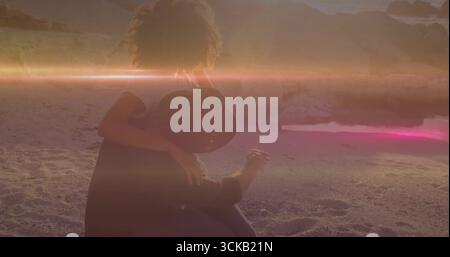 Hugging curly-haired woman and man wearing wide-brimmed hat sitting at water's edge at sunset Stock Photo