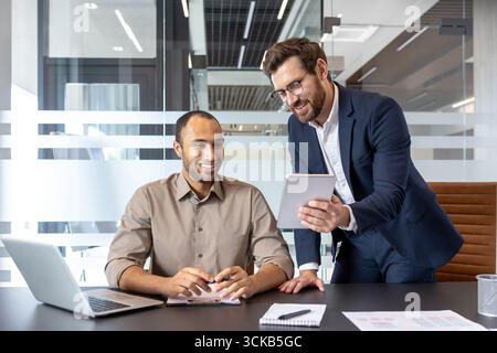 Two smiling male managers discussing a project while analyzing data on a digital tablet and laptop in a bright, modern office environment filled with natural light Stock Photo