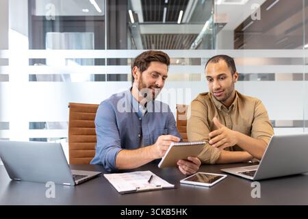 Two smiling male managers are having a productive business meeting, collaborating on a project and discussing notes in a modern office setting, surrounded by laptops and tablets Stock Photo