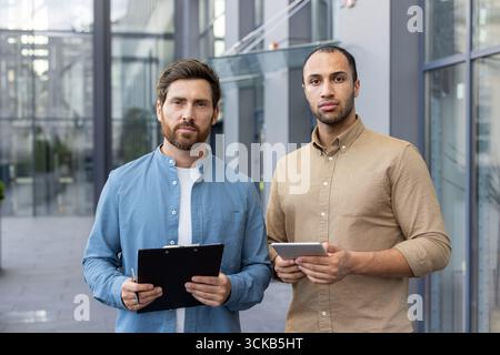 Two businessmen are standing outside modern office building, one holding clipboard and pen, other holding digital tablet, looking serious and professional Stock Photo