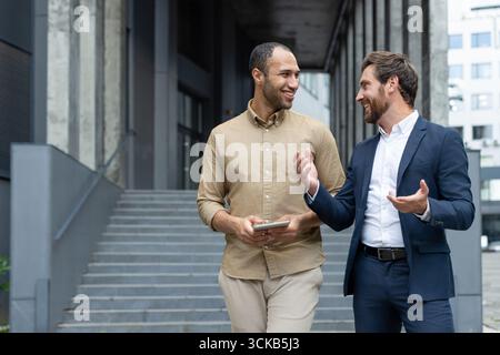 Two businessmen engaging in a friendly conversation while walking outside their office building, discussing work-related topics and collaborating on ideas using a tablet Stock Photo