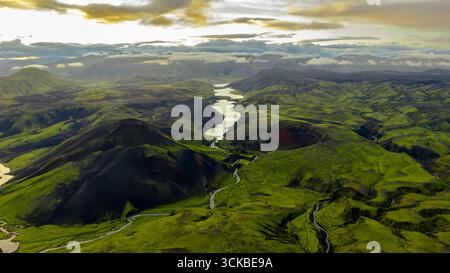 Aerial perspective of Iceland's lush green valleys, dark volcanic hills, a winding river, and a serene lake framed by distant mountains and clouds. Stock Photo