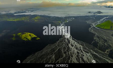 Aerial perspective of Iceland's volcanic plains with braided white rivers, green moss covered hills, and a glacier under low hanging clouds. Stock Photo