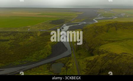 Aerial image of a winding river on a volcanic plain with green fields, a moss covered hill, and a road under an overcast sky in Iceland. Stock Photo